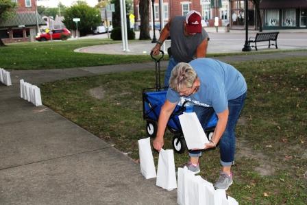 man and woman volunteers place small white paper bags along edge of concrete path beside green grass.
