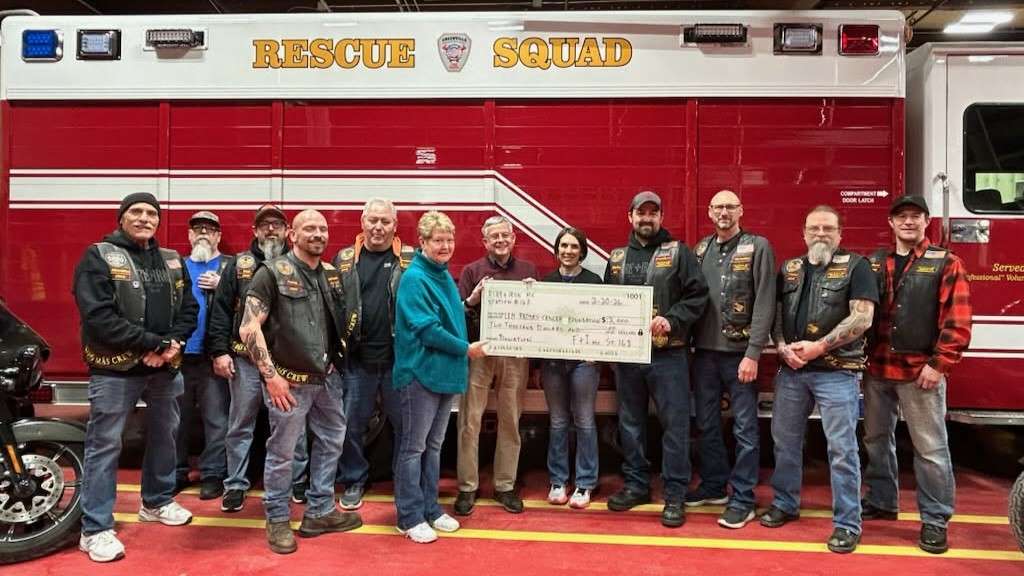 a line of twelve smiling people, many wearing black leather vests with patches, stand in front of a red and white Rescue Squad emergency vehicle posing with an enlarged donation check made out to LRCF