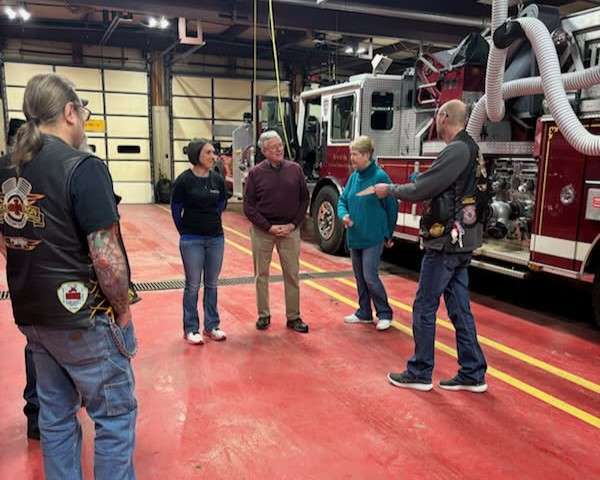 two members of a motorcycle club, wearing black leather vests with patches, have a conversation inside a fire station with three plainclothes representatives of Lem Rhodes Cancer Foundation.