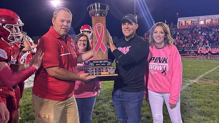 two women in pink bowl sweatshirts and two men in athletic shirts hold up a Pink Bowl trophy with a pink awareness ribbon inside a football stadium