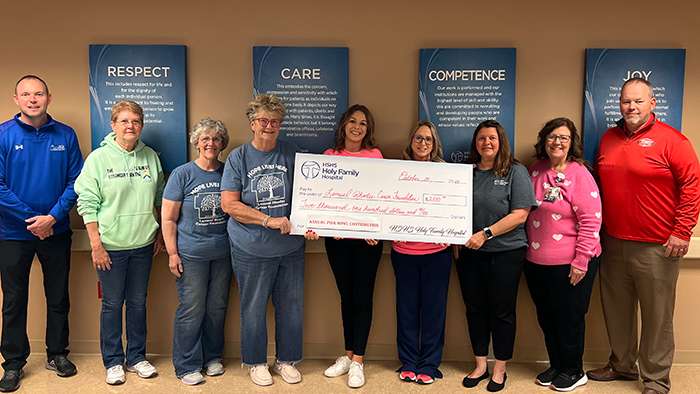 a line of nine smiling people stand in a beige hallway posing with an enlarged onation check made out to LRCF