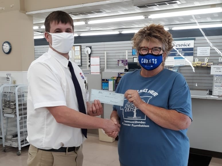 One man and one woman wear fabric face masks shake hands and hold between them a paper donation check.