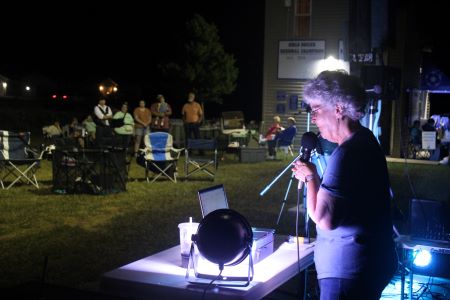 woman in purple shirt with short gray hair speaks into microphone in a spotlight at night in front of people standing and sitting in lawn chairs.