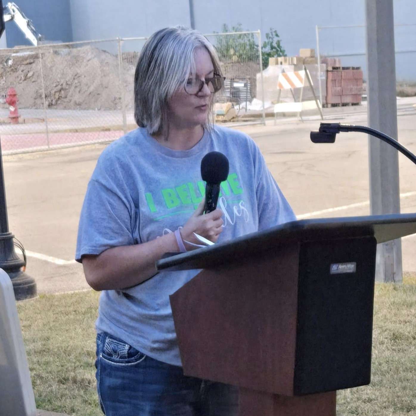 A woman wearing a grey shirt bearing inspirational words stands at a podium, preparing to address the crowd.