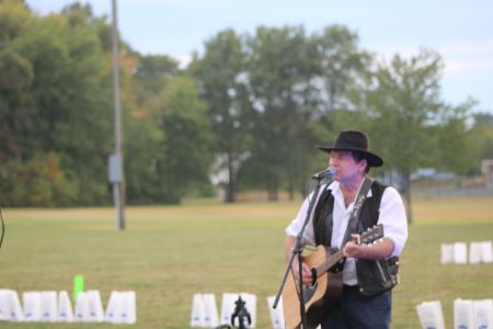 man in white shirt, black vest and black wide brimmed hat sings at microphone while playing brown acoustic guitar outdoors on green athletic field.