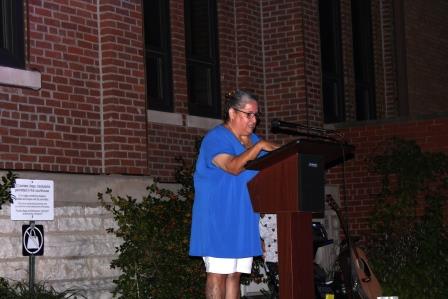 tan gray haired woman wearing long blue shirt stands at microphone and podium in front of large brick building.
