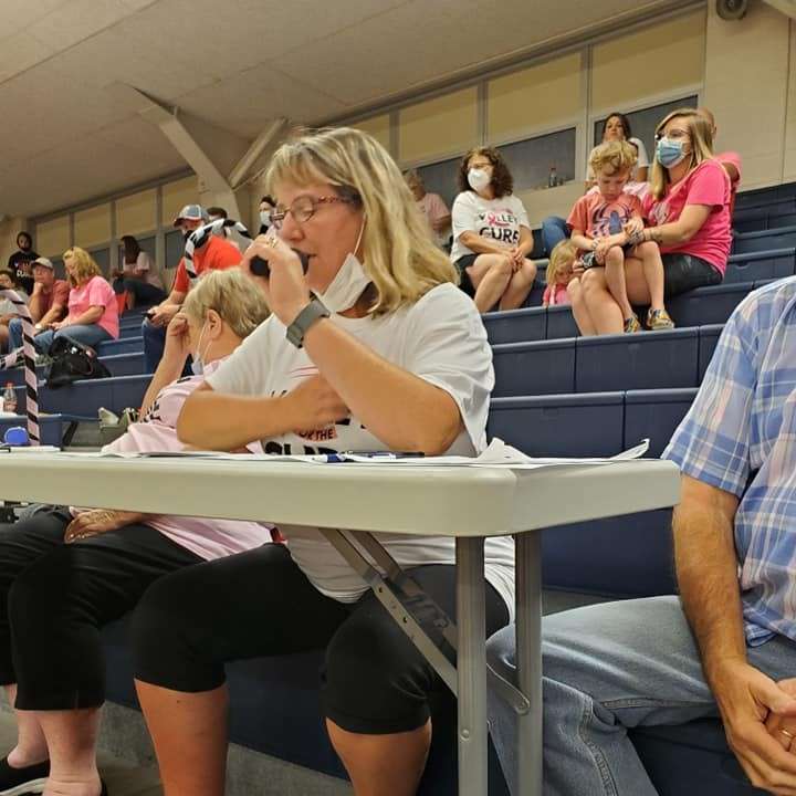 woman in logo tee shirt holds microphone while sitting behind a folding table. behind her are more people sitting on blue school gym bleachers.