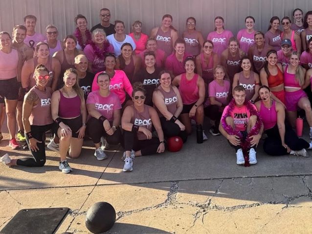 Large, cheerful group of women in pink and black EPOC fitness shirts pose outdoors, with medicine balls and gym equipment in front.