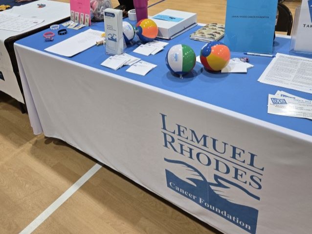 Cancer foundation information table with blue cloth, brochures, rubber bracelets, colorful inflated balls, and a sign reading “Lemuel Rhodes” in a community venue.