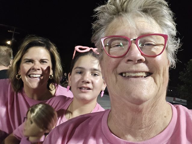 Cheerful group selfie at night: an older woman in pink glasses, a teen, a smiling woman holding a child, all wearing pink shirts.