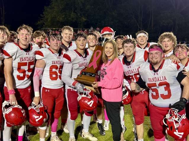 a woman in a pink sweatshirt with a fundraising graphic on the front, presents a wooden trophy to a group of boys wearing red and white high school football uniforms.
