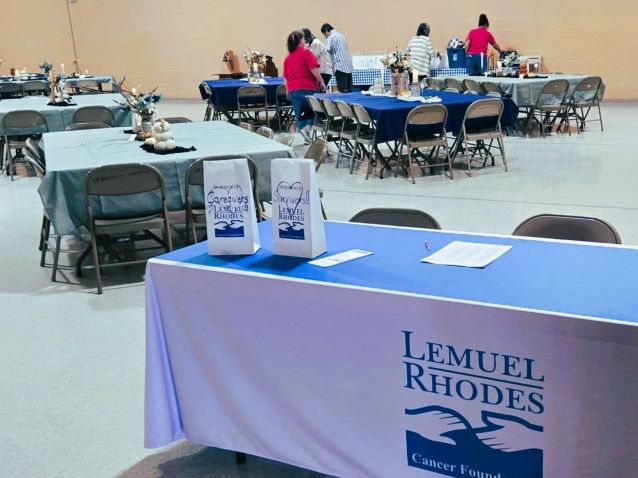 Lemuel Rhodes Cancer Foundation information table in a community hall, with brochures, refreshments, banquet tables, and people gathering.
