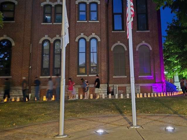 people gather before a three story brick building with flags, illuminated by yellow candles placed in paper bags.