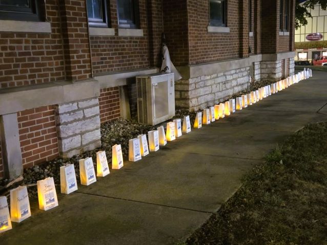 Rows of glowing memorial bags line a brick government building sidewalk at dusk, creating a solemn, peaceful tribute beneath tall arched windows.