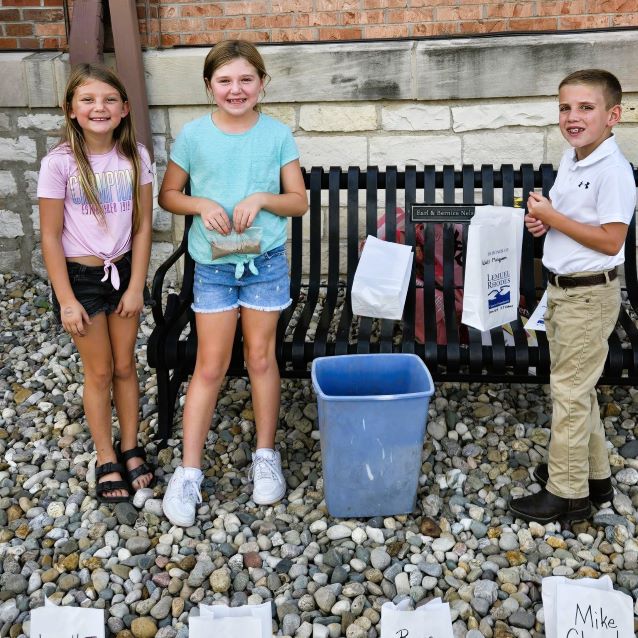 Three smiling children stand by a bench and blue bin outside a brick building, surrounded by labeled luminary memorial bags and colorful river rocks.