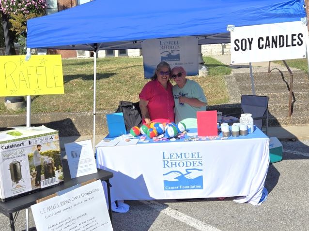 Outdoor craft market booth under a blue canopy, displaying soy candles and colorful cancer awareness goods before a historic brick building.