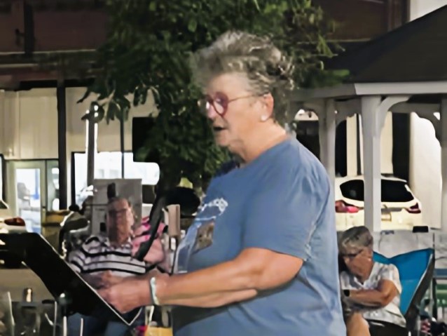 Older woman speaking at a community event, standing behind a “Lemuel Rhodes” sign reading “Uniting Our Community,” with seated people behind her.